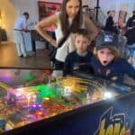 Three children, one looking excited and surprised, play a brightly lit pinball machine in a lively NY living room while an adult woman watches. Other people mingle in the background, creating a fun party atmosphere.