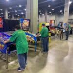 Several people enjoy various pinball machines in a large NY venue with concrete floors and tall pillars. The colorful machines, perfect for party rentals Long Island, feature themes like Street Fighter II and Road Show.