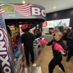 A woman wearing pink gloves punches the Boxing Machine Arcade Game in a store as others watch—ideal party fun, like bounce house or party rentals in Long Island, NY. The machine displays scores and features American flag colors.