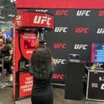 A person with long, wavy hair punches a Boxing Machine Arcade Game at a New York event. UFC logos and a "Test Your Knockout Power" sign are in the background, along with other attendees and party rentals from Long Island.