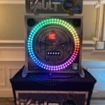 A circular game machine called “The Vault” features a ring of colorful LED lights, a timer set to 30 seconds, and a control panel with buttons, making it a standout choice for party rentals Long Island on a black table against a decorated wall.