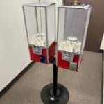 Two empty red and silver candy or capsule vending machines on a black stand, placed in a corner near a brown door in a carpeted indoor area—perfect for party rentals Long Island or bounce house Long Island events in NY.