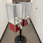 Two red and silver candy or toy vending machines are mounted on a black stand in a carpeted hallway with white walls and a brown door—perfect for adding fun to your next event with party rentals Long Island.