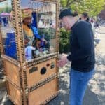 A man in a black shirt and jeans stands in front of a Zoltar fortune-telling machine outdoors, appearing to insert money into the slot. Trees and people hint at a fair or festival, possibly featuring party rentals Long Island style.