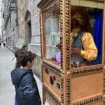 A boy and a white dog stand on a NY city sidewalk in front of a Zoltar fortune teller machine, featuring a mannequin dressed in a yellow shirt and turban inside.