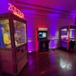 A dimly lit room in NY with a Zoltar fortune teller machine, an NBA Jam arcade game, and a claw machine—all glowing with neon lights against white paneled walls and a polished wood floor—perfect for party rentals Long Island.