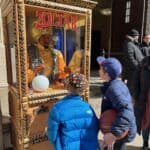 Two children stand in front of a Zoltar fortune-telling machine outside a building in NY. One wears a blue jacket and colorful cap, the other sports a dark jacket and baseball cap, holding a basketball. Several adults are in the background.
