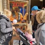People gather around a Zoltar fortune-telling machine outside in NY, including a woman with a stroller, a child, and two adults, as sunlight illuminates the scene—perfect for events with party rentals Long Island.