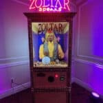 A Zoltar fortune-telling machine with a mannequin dressed in a turban and gold vest, illuminated by purple lights, stands in the corner of a party rentals Long Island showroom under a neon "Zoltar Speaks" sign.