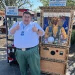 A smiling man in green pants and a white shirt gives two thumbs up while standing next to a Zoltar fortune teller machine outdoors in NY. Signs above advertise tickets and plays for $1.00. Trees, another machine, and party rentals Long Island are visible in the background.