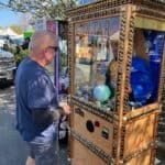 A man in sunglasses and a blue shirt stands in front of a Zoltar fortune-telling machine at an outdoor NY event, holding a drink. Trees, booths, and party rentals Long Island, including a parked vehicle, are visible in the background.