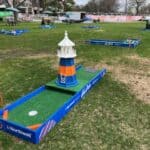 A mini-golf course set up outdoors on grass, featuring blue-bordered holes with fun obstacles and a central lighthouse structure—perfect for party rentals Long Island. Tables and trees are visible in the background.