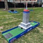 A mini-golf hole designed to look like the Empire State Building stands on a grassy area, perfect for party rentals Long Island fun, with other mini-golf holes and a brick building visible in the background.