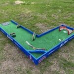 A mini-golf course shaped like an L, with green turf, hockey sticks as obstacles, a small orange goal net, and blue side walls—perfect for outdoor fun on grass. Great for party rentals Long Island events!.
