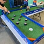 A person assembles a tabletop hockey game with faux turf, pucks, and blue boards labeled "The Park" and "UBS Arena." Tools and construction materials are scattered nearby, just like at top party rentals Long Island setups.