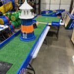 A mini-golf course with green turf and a blue and orange lighthouse obstacle is set up indoors on folding tables, perfect for NY events or party rentals Long Island, surrounded by colorful equipment and storage racks.