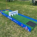 A small outdoor mini-golf course hole in NY features artificial turf, blue borders, orange accents, and a Northwell sign with three arches. The number 6 is marked on the side. Picnic tables are visible—perfect for party rentals Long Island events.