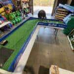A person sets up an L-shaped mini-golf course with artificial grass and blue barriers inside a warehouse in NY, suggesting it's part of party rentals Long Island, with equipment and supplies visible in the background.