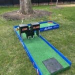 A mini-golf hole with green turf, blue borders, and two black box obstacles labeled "LONG ISLAND"—perfect for party rentals Long Island, NY. The setup is outdoors on grass near a large tree and black metal fence.