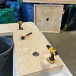 Two wooden cornhole boards with circular holes are being assembled in a workshop, perfect for party rentals Long Island. Drills and a rubber mallet rest on the boards, surrounded by supplies, bags, and a nearby trash bin.