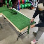 Two people cut and staple green artificial turf onto a rectangular wooden board on a table in a workshop, with colorful cones and party rentals Long Island supplies stacked in the background.
