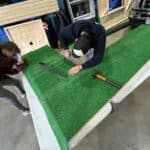 Two people are working on attaching artificial turf to a large triangular board on a table in a workshop, surrounded by tools like a hammer and measuring tape, possibly preparing decor for party rentals Long Island.
