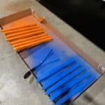 Wooden slats partially spray painted in orange and blue are arranged in a row inside a cardboard box on a concrete floor, reminiscent of creative setups seen at NY party rentals and bounce house Long Island events. The colors blend slightly where the paint overlaps.
