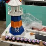 A model lighthouse with blue, orange, and white colors sits on a table next to nine New York Islanders hockey pucks and various crafting materials—perfect inspiration for any bounce house Long Island or party rentals Long Island, NY themed event.