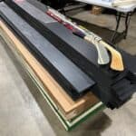 A stack of black foam boards and cardboard rests on the floor, with three hockey sticks placed on top. Nearby, a table holds tools and supplies in a workshop—perfect for prepping for bounce house Long Island party rentals, NY.