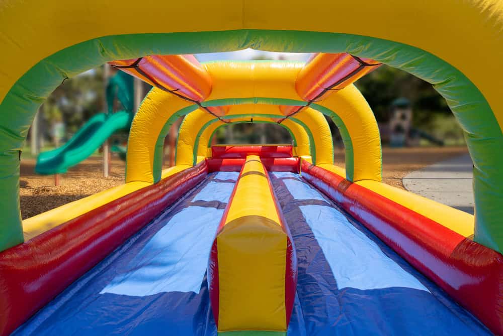 A colorful inflatable obstacle course with yellow, red, and blue sections is set up outdoors in a playground, perfect for party rentals Long Island, with green slides and trees visible in the background.