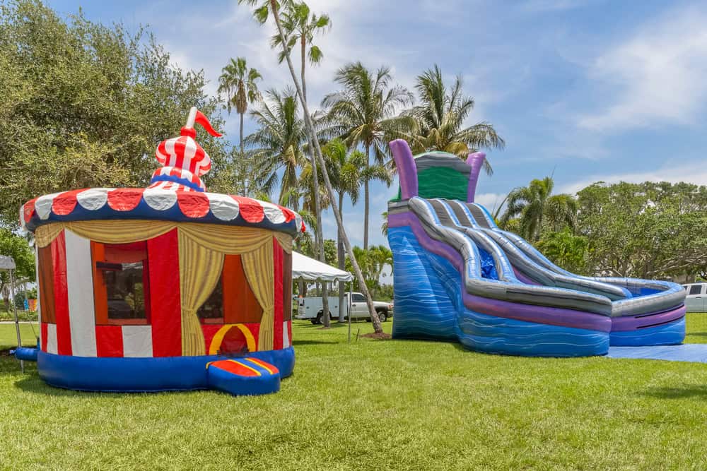 A colorful inflatable circus tent and a large blue inflatable water slide set up on a grassy area with palm trees and a blue sky—perfect for NY parties. Discover top-notch bounce house Long Island rentals for your next event!.