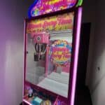 A colorful "Sweet Shoppe" claw machine with bright pink lights and candy-themed decorations stands in a corner at a NY party rentals Long Island event. The sign reads "Winner Every Time," inviting guests to grab fun prizes with the claw.