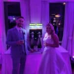 A bride in a white gown and a groom in a light blue suit stand smiling with drinks beside frozen drink machines at their NY wedding reception, featuring purple lighting and fun party rentals Long Island couples love.