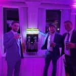 Three men in suits stand smiling around a slushie machine at a party, each holding a drink cup. The room is lit with purple and pink lighting, creating a festive NY atmosphere—perfect for party rentals Long Island style.