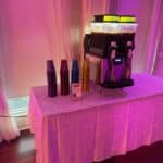A table covered with a white cloth holds a slushie machine and stacks of colorful plastic cups, set against a window with sheer curtains and pink-purple lighting—perfect for party rentals Long Island, NY or your next bounce house Long Island event.