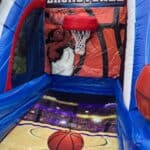 An inflatable basketball game from party rentals Long Island features a cartoon player dunking, a hoop, and a court design with a basketball on the ground. The word "BASKETBALL" is displayed above the hoop. Perfect for NY events!.