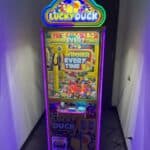 A brightly lit "Lucky Duck" claw machine, perfect for party rentals Long Island, stands in a narrow hallway filled with colorful plastic balls. Plugged into a wall outlet, its sign on top reads “Winner Every Time.”.