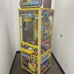 A "Lucky Duck" claw machine filled with colorful rubber ducks sits in a narrow, white-walled hallway with a brown door behind it—just the kind of fun you'd find at NY party rentals Long Island events.