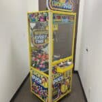 A yellow "Lucky Duck" claw machine filled with colorful rubber ducks stands in a narrow hallway with white walls and a brown door behind it. Perfect for party rentals Long Island, this fun game displays signs reading "Winner Every Time.