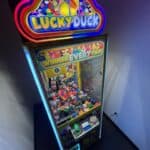 A brightly lit "Lucky Duck" claw machine filled with colorful rubber ducks and plush toys stands in the corner of a dimly lit room at a party rentals Long Island event, with a sign reading "Winner Every Time!" above the toy-filled chamber.