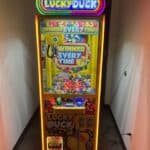 A brightly lit "Lucky Duck" claw machine stands in a hallway, perfect for party rentals Long Island. Filled with colorful toy ducks, the glowing yellow machine promises “Winner Every Time.” A cord is plugged into the wall nearby.