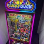 A brightly lit "Lucky Duck" claw machine filled with colorful rubber ducks and toys, perfect for NY party rentals Long Island events, featuring neon lights and a "WINNER EVERY TIME" sign, standing in a dimly lit room.