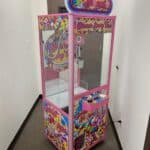 A colorful claw machine labeled "Sweet Shoppe," perfect for party rentals Long Island, features candy-themed decorations in a narrow hallway near a brown door. The pink-framed machine has glass panels, a joystick, and a prize chute at the front.