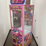 A colorful "Sweet Shoppe" candy claw machine with bright pink trim and vibrant candy-themed decorations stands in a narrow hallway near a brown door—perfect for party rentals Long Island, NY to liven up any event.