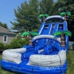 An inflatable blue water slide with palm tree decorations is set up on a grassy lawn, perfect for NY parties. Surrounded by trees and a house under a clear blue sky, it's ideal for party rentals Long Island fun.
