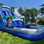 A large blue inflatable water slide with palm tree decorations, perfect for party rentals Long Island, set up on a grassy lawn near a house, with trees and a pool in the background on a sunny NY day.