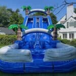 A large, inflatable blue water slide with palm tree decorations is set up on a green lawn in front of a white house on a sunny day—perfect for party rentals Long Island, NY.