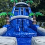A large inflatable blue water slide with two lanes, palm tree decorations, and a center ladder, perfect for party rentals Long Island, NY—set up outdoors on a sunny day with lush green trees in the background.