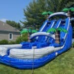 A large blue inflatable water slide with palm tree decorations from party rentals Long Island is set up on a green lawn in a backyard, next to a gray house and surrounded by trees and bushes.