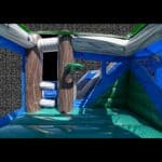 The 7 in 1 Tropical Combo bounce house features blue and green walls, a shiny floor, and tropical-themed trees and logs inside. Shown from one corner against a black background—ideal for party rentals on Long Island, NY.